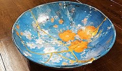 A small pottery bowl is painted light blue with orange and white flowers. Cracks are visible throughout the pottery.