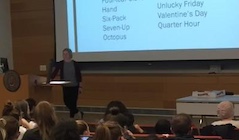 A person standing at a lectern with students in the audience looking at the project screen.