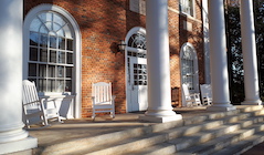 A brick campus building with white empty rocking chairs and white columns.