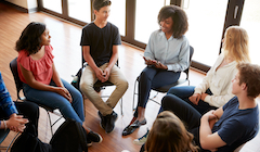 A group of seven diverse students sit in chairs arranged in a circle. One student is talking while the others are actively listening. 