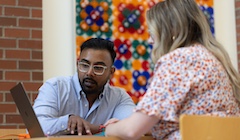 A man and a woman sit at a table looking at a laptop.