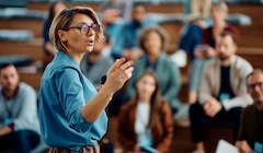 A woman is speaking and instructing in front of a group of students in a lecture hall.