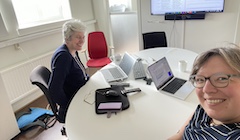 The two authors look at the camera while sitting at a table with computers and papers